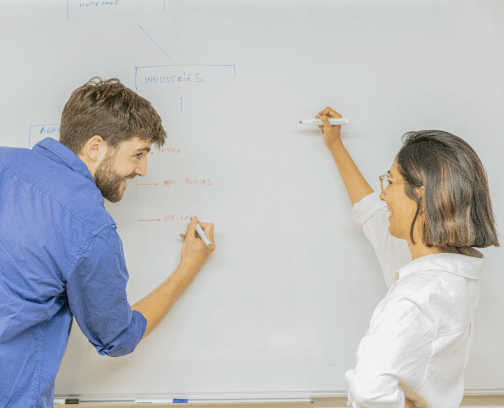 A man with a beard and a blue shirt with rolled up sleeves and a woman with short hair, a white shirt with rolled up sleeves drawing on a white board and smiling at each other.