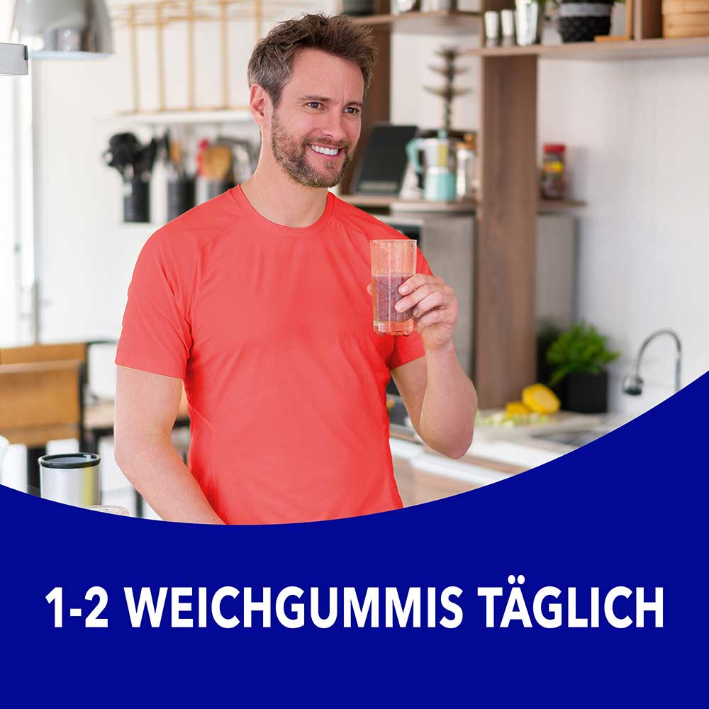 Mann in orangenem Shirt steht in der Küche und hält ein Glas mit dem Text „1-2 Weichgummis täglich"