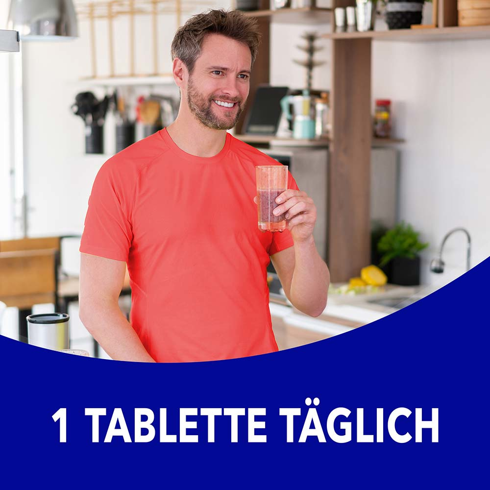 Mann in orangenem Shirt steht in der Küche und hält ein Glas mit dem Text „1 Tablette täglich"