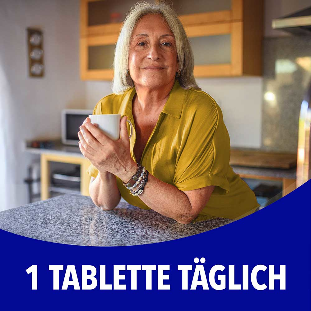 Ältere Frau sitzt entspannt in der Küche und hält eine Tasse, mit dem Text „1 Tablette täglich"