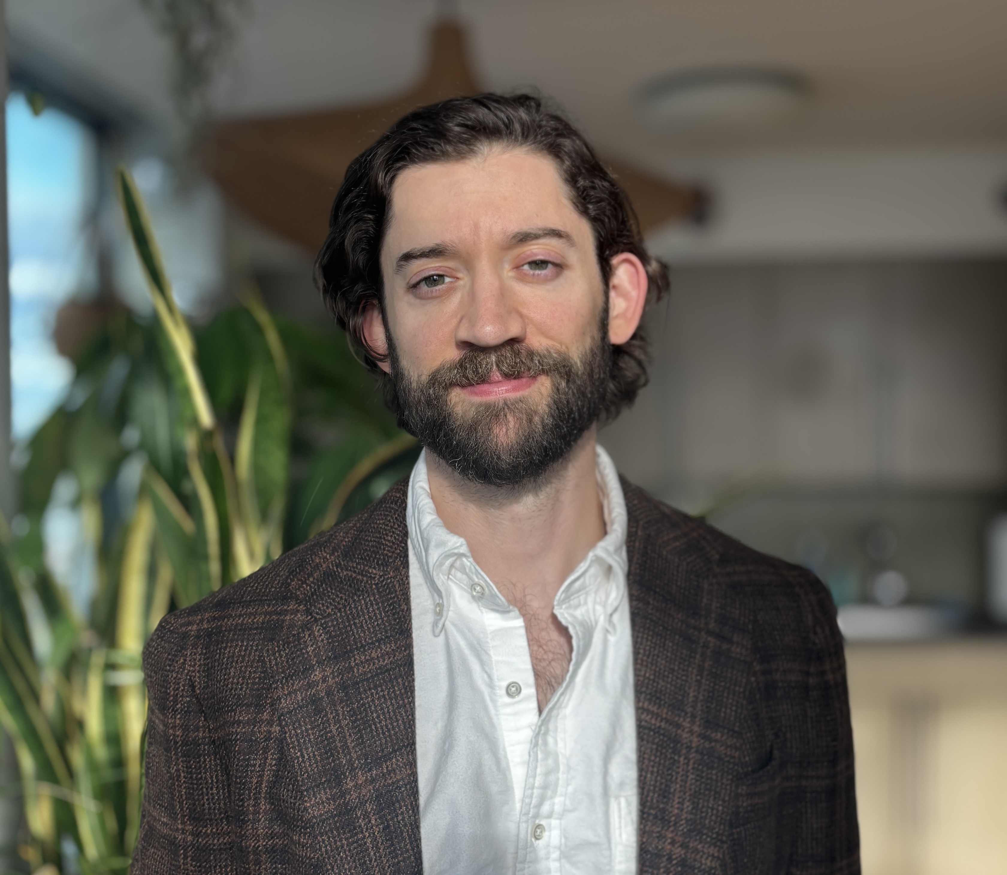 Photo of a white man with dark brown hair, mustache, and beard, wearing a brown suit jacket and white dress shirt