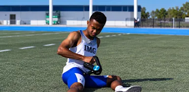 An athlete sits on a field using a handheld massage device on his leg.