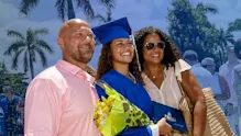 IMG Academy graduate in blue cap and gown posing with family during graduation ceremony, holding flowers and a diploma outdoors with palm trees in the background.