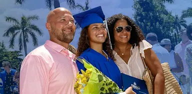 IMG Academy graduate in blue cap and gown posing with family during graduation ceremony, holding flowers and a diploma outdoors with palm trees in the background.