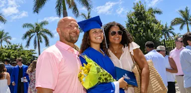 A graduate in a blue cap and gown poses with her family while holding flowers and a diploma folder.