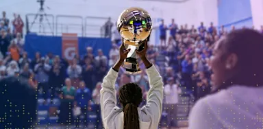 IMG Academy student-athlete holds a championship trophy overhead in a packed gymnasium as teammates and fans celebrate behind her.