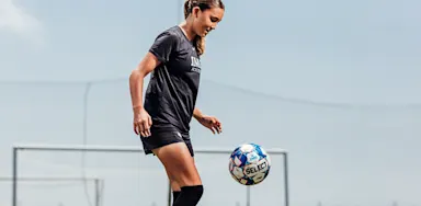An IMG Academy soccer player balances a ball in the air while practicing control, standing on a field in a black training outfit with a goal visible in the background.