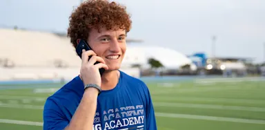 A smiling student-athlete talks on a phone beside a field.