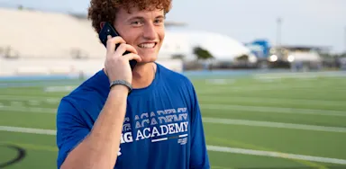 Male IMG Academy student-athlete stands on an outdoor athletic field, smiling while holding a phone to his ear. He wears a blue athletic shirt and a wristband on his left arm.
