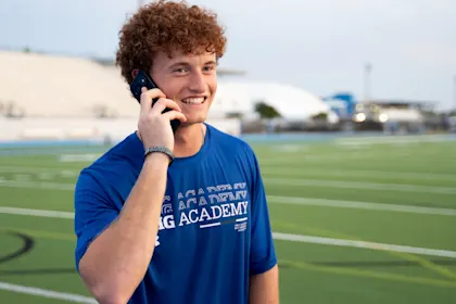 Male IMG Academy student-athlete stands on an outdoor athletic field, smiling while holding a phone to his ear. He wears a blue athletic shirt and a wristband on his left arm.