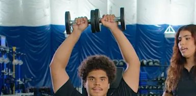 An athlete holds dumbbells overhead while a trainer observes.