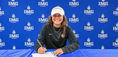 A smiling student-athlete signs paperwork at a table in front of an IMG Academy backdrop.
