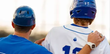 A baseball coach with his arm around a player's shoulder, viewed from behind, during a game at sunset.