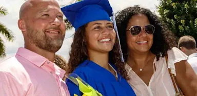 A young woman in a bright blue cap and gown poses with her family at an outdoor graduation ceremony, holding a bouquet of yellow flowers. Palm trees and other smiling attendees are visible in the sunny background.