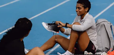 A seated athlete adjusts her shoe beside a blue running track.