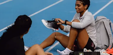 A female athlete sits on a blue track tying her running shoe while talking with a teammate, with a backpack and training gear beside her.