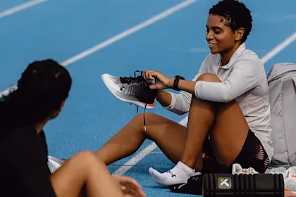 A female athlete sits on a blue track tying her running shoe while talking with a teammate, with a backpack and training gear beside her.