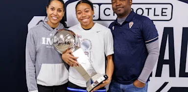 Championship winner (student-athlete) with trophy, flanked by two proud adults (coaches/parents), celebrating at the Chipotle Nationals basketball tournament.