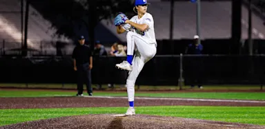High school baseball player in full white uniform winds up to pitch on a lit field at night. He stands tall on the mound with focused posture, ready to deliver the ball.