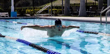 An IMG Academy student-athlete swimmer performs the butterfly stroke in an outdoor pool, with water splashing as their arms extend across the lane.