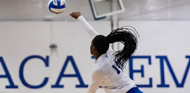 An IMG Academy student-athlete volleyball player jumps to serve the ball in an indoor gym, striking it overhead with a powerful motion.