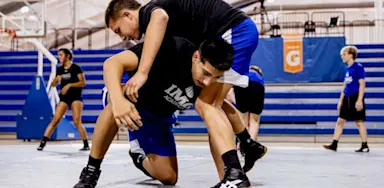 Two IMG Academy athletes practice wrestling in a gym, with one lifting the other onto his shoulders while kneeling on a mat. Other athletes train in the background near blue bleachers and gym equipment.