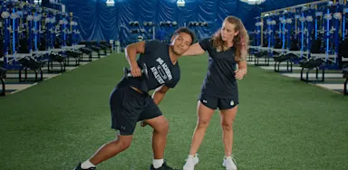 A trainer guides an athlete through a standing exercise on indoor turf.