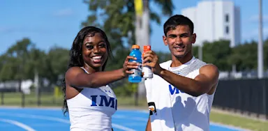 Two athletes smile while holding sports-drink bottles together on a field.