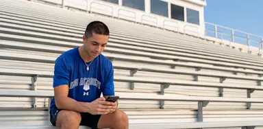 A student-athlete looks at his phone while seated on outdoor bleachers.