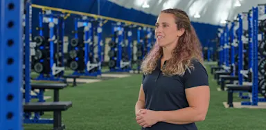 A trainer stands beside weight racks with her hands clasped.