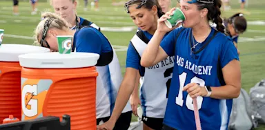 Female lacrosse athletes taking a hydration break, drinking from cups near orange Gatorade coolers on the sideline of a field.