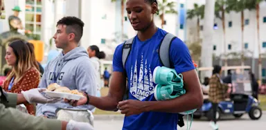 IMG Academy student-athlete in blue shirt and backpack receives a plate of food from a server, he is surrounded by palm trees and students in the background.