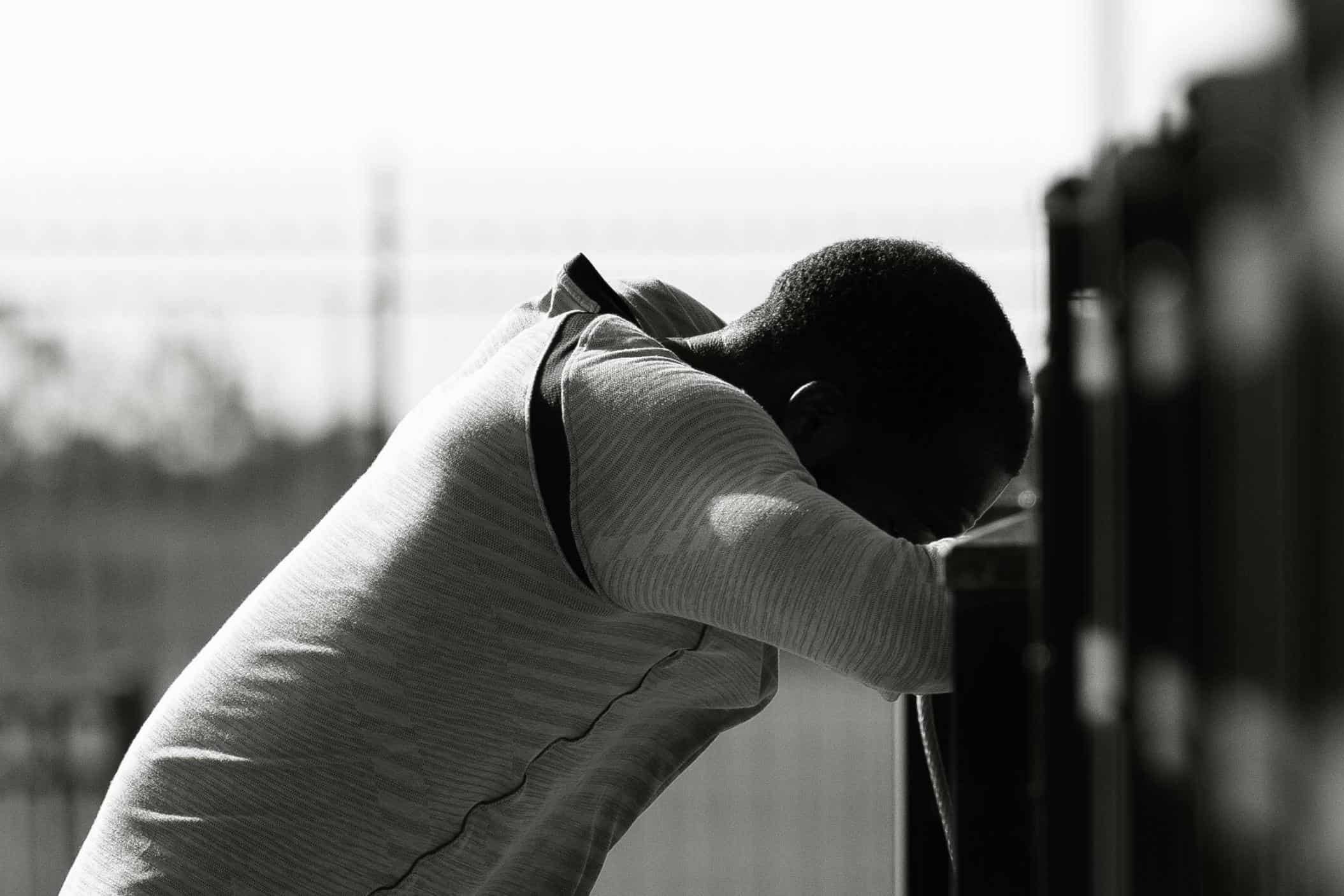 A black and white photo of a person leaning forward against a locker or railing, head down and arms crossed in front. The posture and lighting evoke a mood of exhaustion, stress, or contemplation, highlighting the emotional weight often carried by student-athletes.