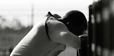 A black and white photo of a person leaning forward against a locker or railing, head down and arms crossed in front. The posture and lighting evoke a mood of exhaustion, stress, or contemplation, highlighting the emotional weight often carried by student-athletes.