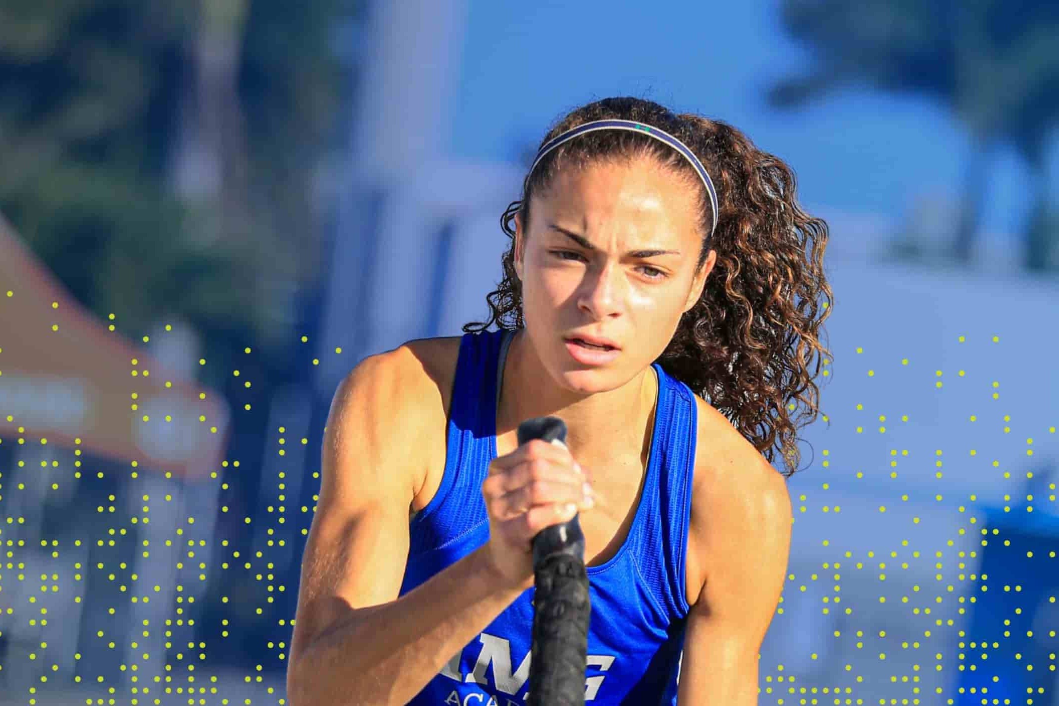 A focused student-athlete in a blue IMG Academy tank top works out with battle ropes during an outdoor training session, with determination visible on her face and palm trees in the blurred background.