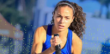 A focused student-athlete in a blue IMG Academy tank top works out with battle ropes during an outdoor training session, with determination visible on her face and palm trees in the blurred background.