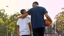 Parent walks with a young athlete on an outdoor sports field, arm around the athlete’s shoulders while holding a soccer ball, symbolizing mentorship, guidance, and support.