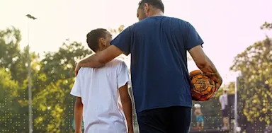 Parent walks with a young athlete on an outdoor sports field, arm around the athlete’s shoulders while holding a soccer ball, symbolizing mentorship, guidance, and support.