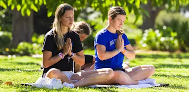 Students sit cross-legged on yoga mats outdoors with their hands together in front of their chests.