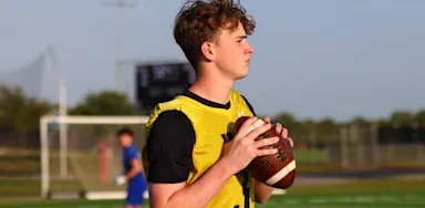 An IMG Academy student-athlete quarterback holds a football in a ready position on the field, looking downfield with a goalpost and other athletes in the background.