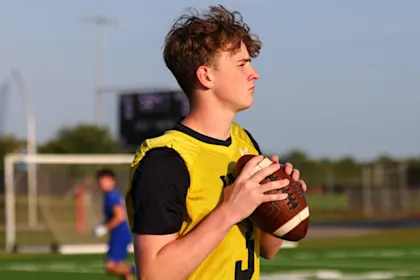 An IMG Academy student-athlete quarterback holds a football in a ready position on the field, looking downfield with a goalpost and other athletes in the background.