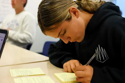 Student-athlete wearing a black sweatshirt taking notes or journaling on yellow paper reflecting on past athletic performance.