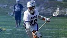 An IMG Academy lacrosse player in full protective gear runs across the field while cradling the ball in his stick, focused ahead during practice, with a teammate visible in the background.