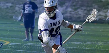 An IMG Academy lacrosse player in full protective gear runs across the field while cradling the ball in his stick, focused ahead during practice, with a teammate visible in the background.