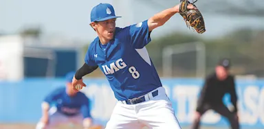 A IMG Academy baseball pitcher in uniform winds up to throw the ball on the field, with teammates and an umpire visible in the background.
