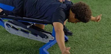 An athlete lies face down on a bench with his arms extended during a shoulder exercise.