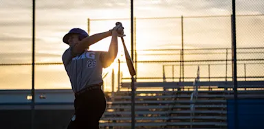A baseball player follows through on a swing in evening sunlight.
