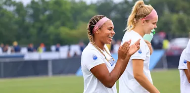 Two soccer players walk across a field, with one smiling and applauding.