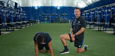 A trainer kneels beside an athlete performing a wrist-mobility exercise on turf.