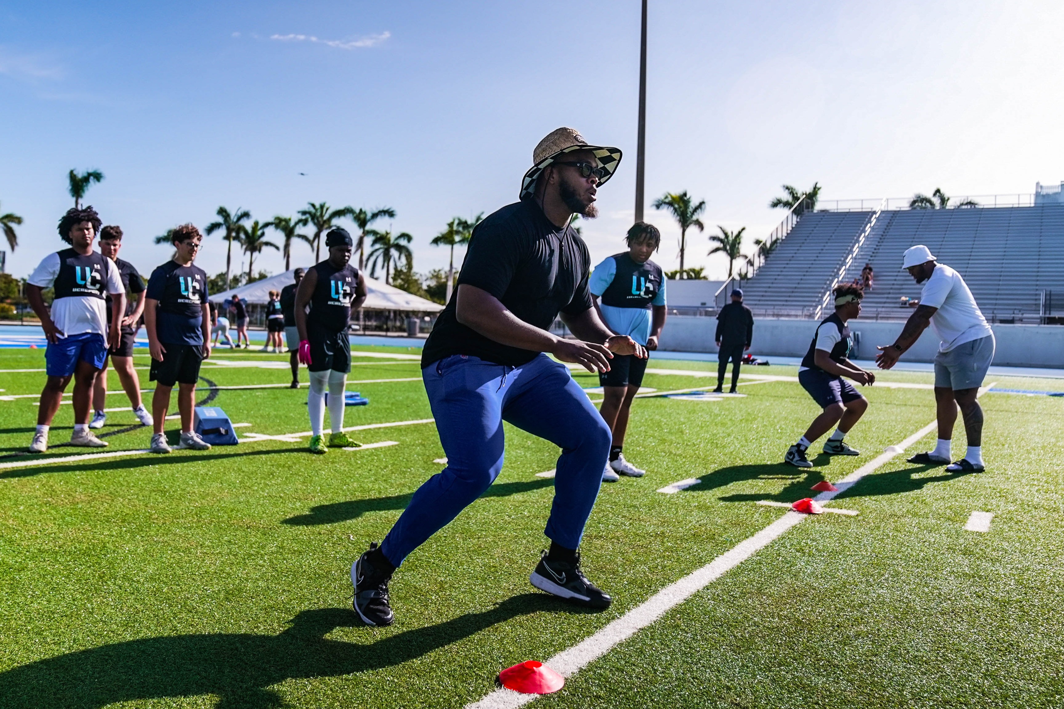 A football coach in a sun hat leads student-athletes through cone drills on a bright green turf field, while others watch and wait their turn during outdoor training at IMG Academy.